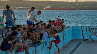 People sit along the edge of an abandoned swimming pool across from a tanker terminal along the port