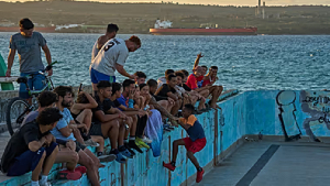 People sit along the edge of an abandoned swimming pool across from a tanker terminal along the port