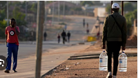 A man carrying water bottles walks down a dusty street in Hamanskraal