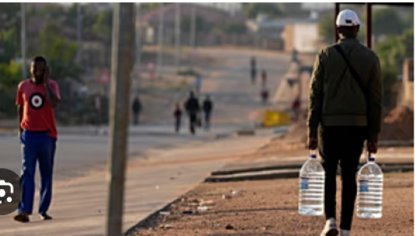 A man carrying water bottles walks down a dusty street in Hamanskraal