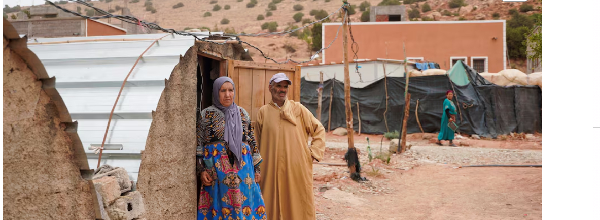 Residents stand outside their NGO-built makeshift shelter with a tin roof