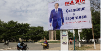 Motorcyclists ride by an election campaign poster for the incumbent President Paul Biya in Maroua