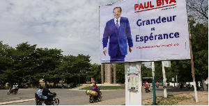 Motorcyclists ride by an election campaign poster for the incumbent President Paul Biya in Maroua