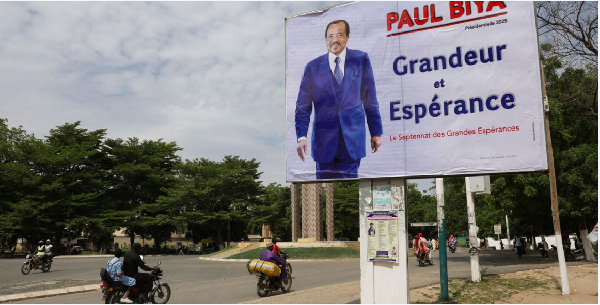 Motorcyclists ride by an election campaign poster for the incumbent President Paul Biya in Maroua