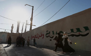 Syrian women walk past a wall with Arabic words
