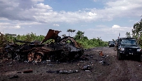 Cars passing by a destroyed military vehicle in Rugari