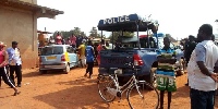 The photo shows a police vehicle with several onlookers gathered at a crime scene